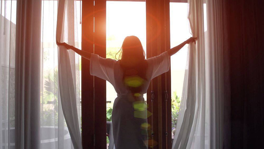 Young woman in bathrobe open curtains and stretch standing near the window at home.